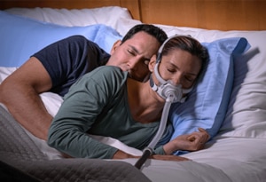 A women sleeping peacefully on her side wearing the ResMed AirFit F40 CPAP mask with her partner cuddled up behind her.