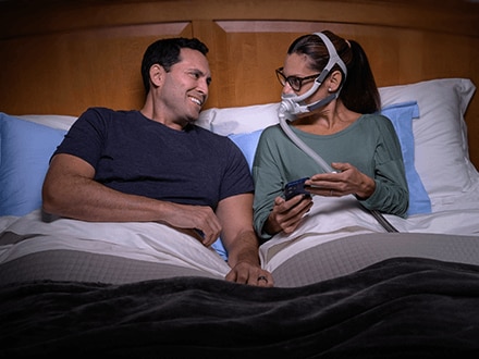 A woman wearing a ResMed AirFit F40 full face Minimalist mask while sitting up in bed, showing her partner something on her phone.