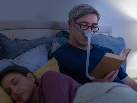 A man with glasses reading in bed while wearing a ResMed AirFit N30 nasal Minimalist mask, as his wife sleeps beside him.