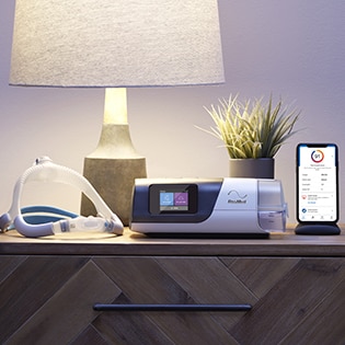 An Air11 sleep apnoea device, mask and a mobile phone showing myAir app on a bedside table.