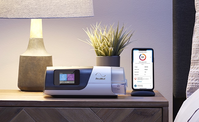 An Air11 sleep apnoea device, mask and a mobile phone showing myAir app on a bedside table.