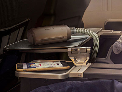 An aeroplane tray table with a AirMini portable CPAP device resting on it, alongside a mobile phone showing the AirMini app on its screen.
