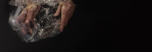 A pair of hands plunging a CPAP device humidifier tub into water against a black background.