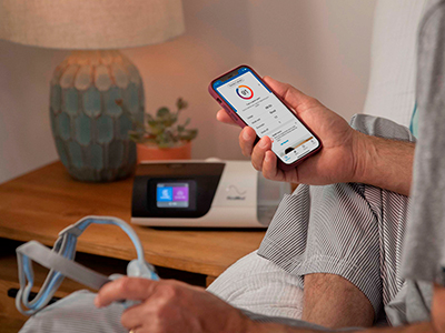 Someone sitting in bed holding a smartphone showing the myAir app in one hand, with a mask in the other, and a therapy device on the bedside table