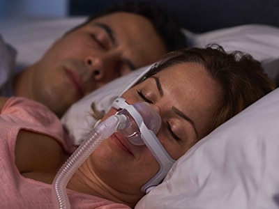 A close-up of a woman sleeping soundly while wearing a CPAP mask, with her partner asleep in the background.