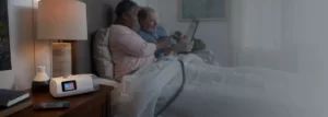 A couple in bed looking at an electronic tablet. The woman has an AirCurve 11 ASV PaceWave machine mask on her lap. The mask is attached to the device on her bedside table.
