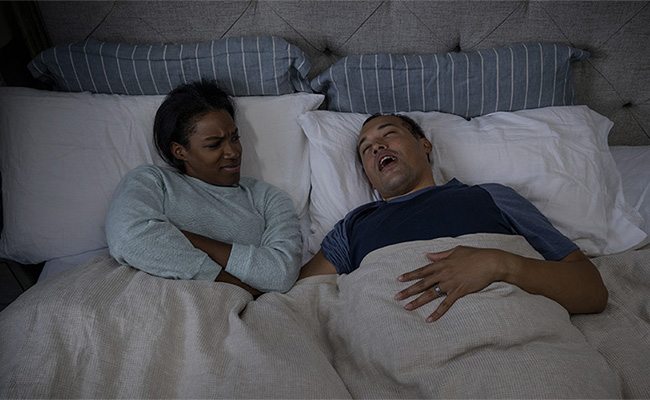 A woman looking at her partner snoring in bed and wondering what is causing his snoring