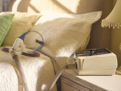 A ResMed ventilator on a bedside table with its mask attached by tubing and resting on the bed.