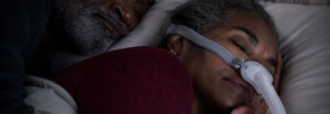 A close-up of a woman sleeping soundly while wearing a CPAP nasal mask, with her partner beside her.