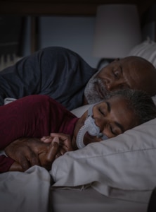A close-up of a woman sleeping soundly while wearing a CPAP nasal mask, with her partner beside her.