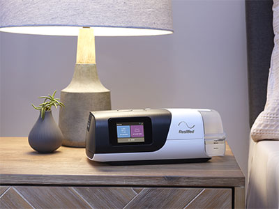 A ResMed CPAP device for treating sleep apnoea on a bedside table next to a lamp.