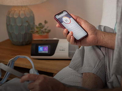 Someone sitting on a bed, holding a mobile phone with the ResMed myAir app on it in one hand and a CPAP mask in the other, with a CPAP device on the bedside table.