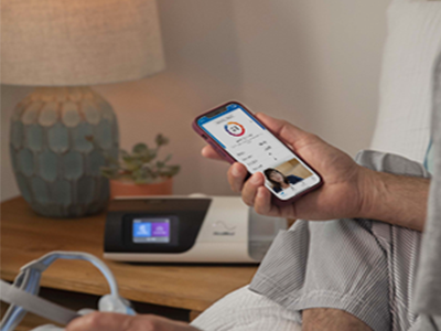 Someone sitting in bed holding a smartphone showing the myAir app in one hand, with a mask in the other, and a therapy device on the bedside table.