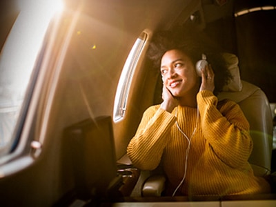 A young woman looking out of a plane window while wearing headphones, with sunlight streaming through the window.