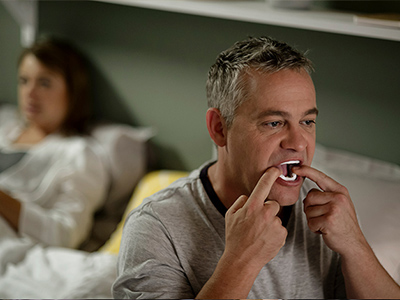 Man sitting on his bed placing a mandibular repositioning device into his mouth before sleep to help prevent snoring.